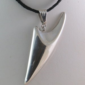 Puede incluir: Un colgante de plata con forma de punta abstracta y afilada, que cuelga de un collar de cordón negro.