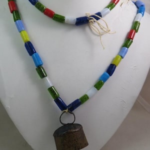 Puede incluir: Un collar colorido con un colgante de campana. El collar presenta una cadena de cuentas cilíndricas multicolores en tonos azules, verdes, rojos, blancos y amarillos. Una pequeña campana de metal envejecido cuelga en el centro.