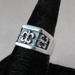 Puede incluir: Un anillo de plata con una banda cuadrada que presenta un diseño repetitivo de lirios estilizados.