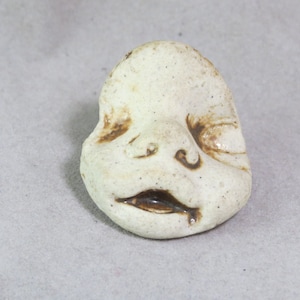 Puede incluir: Una escultura de cerámica blanca de una cara con detalles pintados en marrón. La cara tiene la boca y los ojos cerrados.