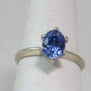May include: A silver ring with a large, oval-shaped, deep blue gemstone. The gemstone is held in place by six prongs, and the band is plain, polished silver. The ring is shown on a white surface.