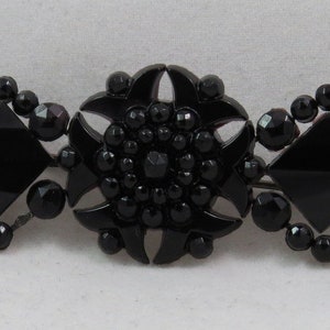 May include: A black glass brooch with a central starburst design surrounded by two square shapes. The brooch is made of black glass beads and has a vintage look.