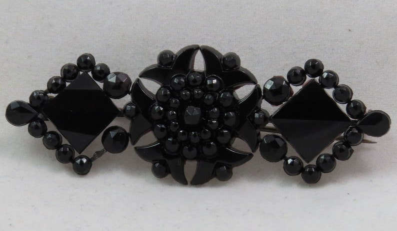 May include: A black glass brooch with a central starburst design surrounded by two square shapes. The brooch is made of black glass beads and has a vintage look.