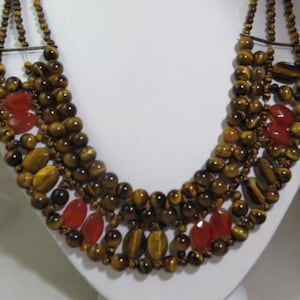 Puede incluir: Collar de múltiples hilos con cuentas de ojo de tigre en tonos marrón y dorado, intercaladas con cuentas ovaladas y redondas. También se incorporan piedras ovaladas de color rojo anaranjado. El collar se exhibe sobre una superficie blanca.