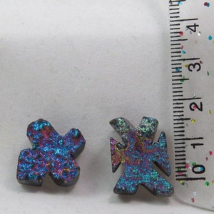 Puede incluir: Dos cruces de piedra preciosa negra iridiscente, una con una forma más redondeada y la otra con una forma más puntiaguda, se muestran junto a una regla. La regla está marcada en pulgadas y muestra que las cruces tienen aproximadamente 1 pulgada de tamaño.