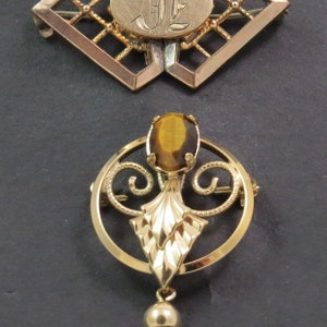 Puede incluir: Dos broches de tono dorado. El primer broche es un marco de forma de diamante con un óvalo en el centro que tiene las iniciales "JE" grabadas. El segundo broche es un marco circular con una piedra preciosa marrón en el centro y una borla dorada que cuelga de la parte inferior.