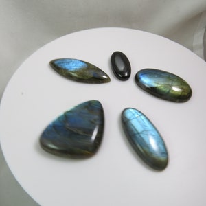 Puede incluir: Cinco cabujones de piedra preciosa pulidos, cuatro son de labradorita azul verdosa iridiscente y uno es negro. Las piedras están dispuestas en forma de estrella sobre una superficie blanca.