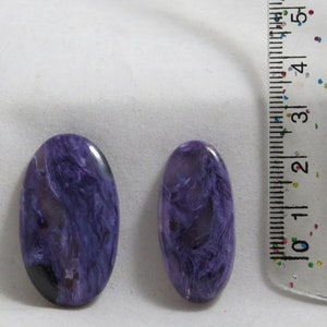 Puede incluir: Dos cabujones ovalados de color púrpura y blanco, cada uno de aproximadamente 2,5 cm de largo, junto a una regla.
