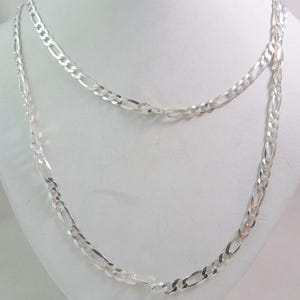 Puede incluir: Un collar de cadena de plata con un patrón de eslabones figaro. La cadena está superpuesta sobre sí misma, creando un aspecto de doble capa.