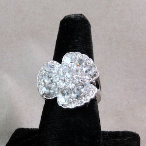 Puede incluir: Un anillo de plata con un gran diseño de flor de cristal transparente. El anillo está en un expositor de anillos negro.