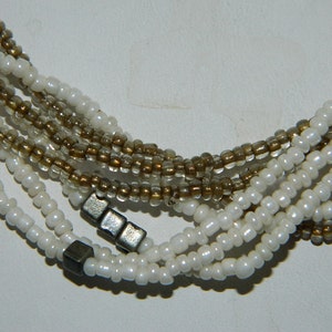 Puede incluir: Un collar de múltiples hilos hecho de cuentas de semillas blancas y marrones con detalles metálicos plateados.
