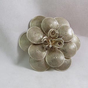 Puede incluir: Un broche de flor de filigrana de plata con un diseño delicado e intrincado. El broche está hecho de muchos círculos pequeños interconectados que crean un patrón floral.
