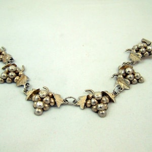 Puede incluir: Un brazalete de plata con un motivo de uva. El brazalete está formado por cinco racimos de uva, cada uno con un racimo de pequeñas cuentas de plata y hojas.