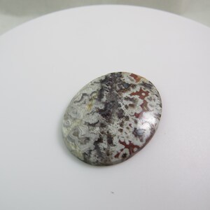 Puede incluir: Un cabujón de ágata blanco, gris y rojo con un patrón de remolino. El cabujón tiene forma ovalada y una superficie lisa y pulida.