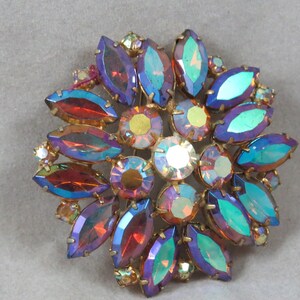 Puede incluir: Un broche grande iridiscente con un diseño de flor. El broche está hecho de metal dorado y presenta una variedad de piedras de imitación de colores de piedras preciosas en tonos de morado, azul, rojo y verde.