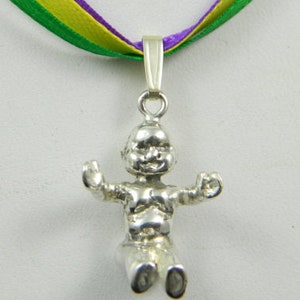 Puede incluir: Collar con dije de plata que presenta una pequeña figura detallada de un bebé sentado con los brazos extendidos. El collar está unido a una cinta verde, amarilla y morada.