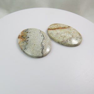 Puede incluir: Dos cabujones de ágata ovalados, pulidos, con un patrón de color blanco, gris y marrón. Los cabujones están sobre una superficie blanca.