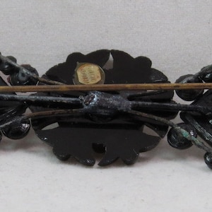 May include: A black and gold brooch with a floral design and a square shape. The brooch has a pin on the back and a label that reads "Made in Austria".