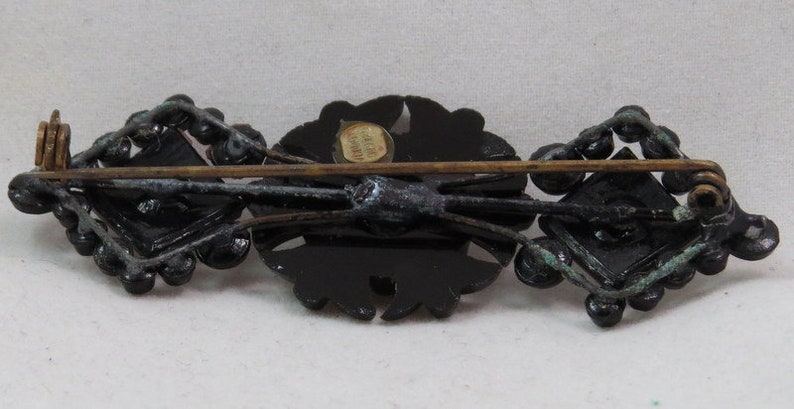 May include: A black and gold brooch with a floral design and a square shape. The brooch has a pin on the back and a label that reads "Made in Austria".