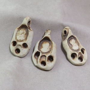 Puede incluir: Tres colgantes de cerámica con un esmalte blanco y detalles marrones. Cada colgante tiene un diseño único con un agujero en el centro y un pequeño agujero en el lateral.
