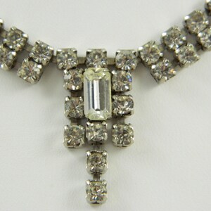 Puede incluir: Un collar de plata con una fila de diamantes de imitación transparentes a cada lado y un diamante de imitación rectangular más grande en el centro. El collar tiene un colgante de gota con un grupo de diamantes de imitación más pequeños.