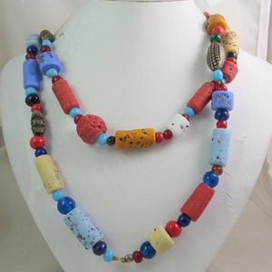 Puede incluir: Un collar de múltiples hilos con cuentas coloridas y texturizadas. Las cuentas son en tonos de rojo, naranja, amarillo, azul y verde. Algunas cuentas son redondas, mientras que otras son cilíndricas.