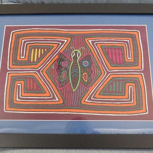 Puede incluir: Una obra de arte enmarcada que presenta una mariposa colorida con patrones geométricos en tonos naranja, verde, rosa y amarillo. La mariposa está rodeada por un diseño intrincado de líneas y formas entrelazadas.