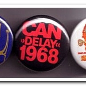 May include: Three round, pinback buttons with a black, white, and blue color scheme. The buttons feature the word "CAN" in white lettering on a blue background, the word "CAN" in red lettering on a black background with the text "Delay 1968", and a red and orange illustration of a person's head with the word "CAN" in white lettering on a white background.