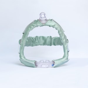 May include: A sage green CPAP headgear strap cover set featuring a smooth, satin-like fabric finish. The assembly includes a top crown piece with a central clear plastic connector and a lower nasal pillow interface with a clear silicone cushion.