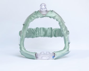 Könnte beinhalten: Ein salbeigrünes CPAP-Kopfband-Abdeckungsset mit einer glatten, satinartigen Stoffoberfläche. Das Set besteht aus einem oberen Kopfteil mit einem zentralen, durchsichtigen Kunststoffverbinder und einem unteren Nasenpolster-Interface.