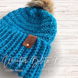 May include: A turquoise blue crochet beanie with a faux fur pom pom on top. The beanie has a brown leather patch with the text "CC Threads" on it. The beanie is on a white wood background. Winter Blythe Beanie Crochet Pattern.