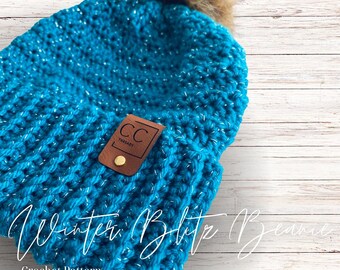May include: A turquoise blue crochet beanie with a faux fur pom pom on top. The beanie has a brown leather patch with the text "CC Threads" on it. The beanie is on a white wood background. Winter Blythe Beanie Crochet Pattern.