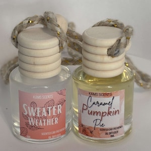 Puede incluir: Dos pequeñas botellas de vidrio con tapas de madera y una cuerda dorada. Las botellas contienen ambientadores para automóviles. Las etiquetas de las botellas dicen "KAMS SCENTS Sweater Weather Scented Car Freshener Oil Diffuser" y "KAMS SCENTS Caramel Pumpkin Pie Scented Car Freshener Oil Diffuser".