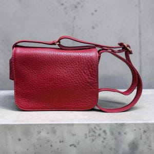 Puede incluir: Un bolso bandolera de cuero rojo con una superficie texturizada. El bolso tiene forma rectangular con esquinas redondeadas y cierre de solapa. Cuenta con una correa larga y ajustable y herrajes dorados.