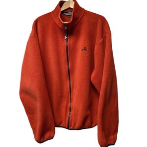 May include: A burnt orange fleece jacket with a full-length black zipper and a high collar. The jacket has a small black logo on the chest and is hanging on a wooden hanger. The fleece material appears soft and warm.
