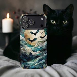 May include: A phone case featuring a stained-glass design with a full moon, bats, and swirling blue and green clouds. The case has a black camera module and a gold-colored border. A black cat is in the background.
