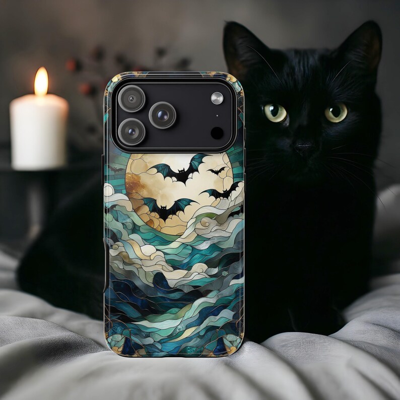 May include: A phone case featuring a stained-glass design with a full moon, bats, and swirling blue and green clouds. The case has a black camera module and a gold-colored border. A black cat is in the background.