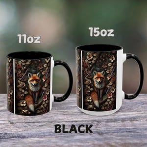Op de afbeelding: Twee zwarte en witte keramische mokken met een vos-illustratie. De mokken zijn verkrijgbaar in de maten 11 oz en 15 oz. De vos is omringd door bloemen en bladeren. De tekst "BLACK" is onder de mokken gedrukt.