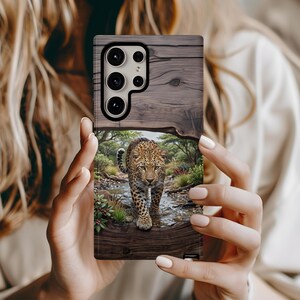 May include: A rectangular smartphone case featuring a printed design of a leopard walking through a shallow stream in a wooded area. The upper portion of the case displays a dark brown wood grain pattern, while the lower section shows the leopard.