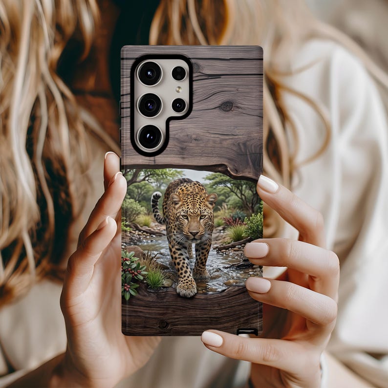 May include: A rectangular smartphone case featuring a printed design of a leopard walking through a shallow stream in a wooded area. The upper portion of the case displays a dark brown wood grain pattern, while the lower section shows the leopard.
