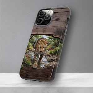 May include: A smartphone case featuring a printed design of a leopard walking through a shallow stream. The background of the case mimics a dark brown wood grain texture, framing the central wildlife scene with green trees and rocky terrain.