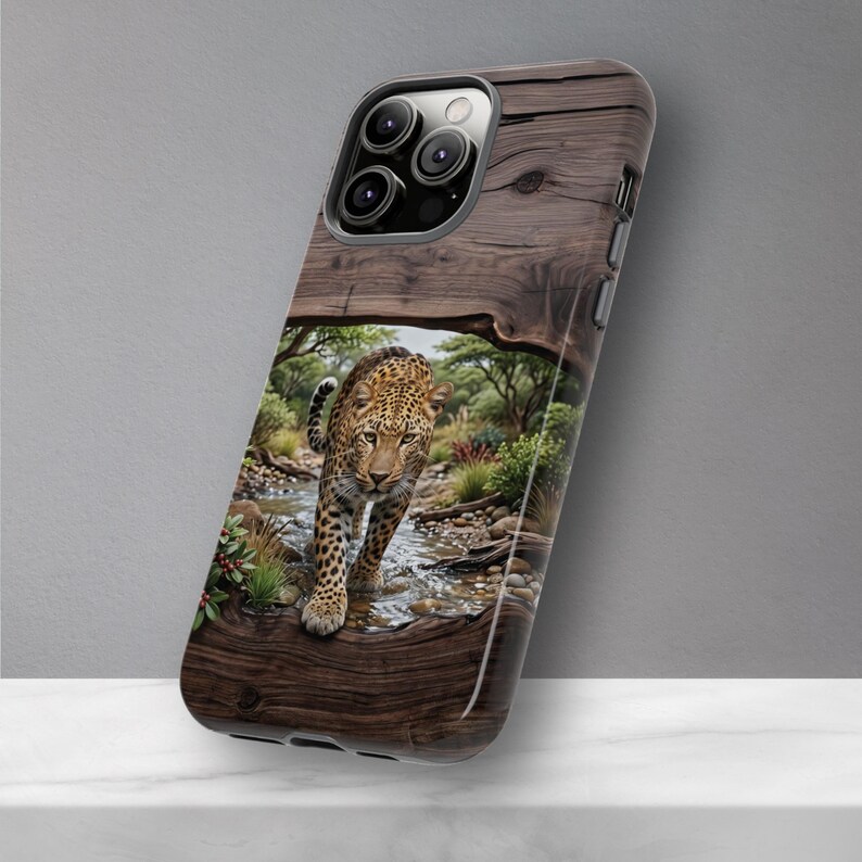 May include: A smartphone case featuring a printed design of a leopard walking through a shallow stream. The background of the case mimics a dark brown wood grain texture, framing the central wildlife scene with green trees and rocky terrain.
