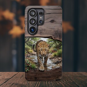 May include: A smartphone case featuring a wood grain texture on the top and bottom sections. The center displays a detailed illustration of a leopard walking through a shallow stream in a natural landscape with green trees and bushes.
