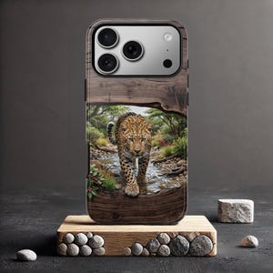May include: A smartphone case featuring a high-resolution print of a leopard walking through a shallow stream in a wooded landscape. The design is framed by a dark wood grain border, and the case includes a cutout for a triple-lens camera system.