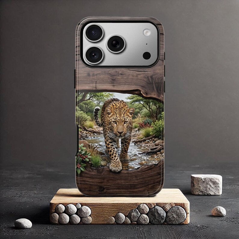 May include: A smartphone case featuring a high-resolution print of a leopard walking through a shallow stream in a wooded landscape. The design is framed by a dark wood grain border, and the case includes a cutout for a triple-lens camera system.