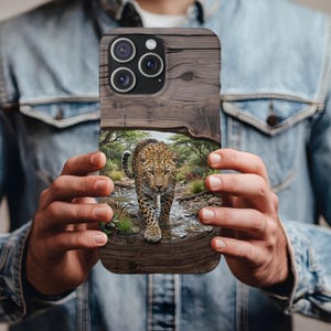 May include: A rectangular smartphone case featuring a wood grain texture on the top and bottom sections. The center displays a detailed graphic of a leopard walking through a shallow stream in a grassy, wooded environment with green foliage.