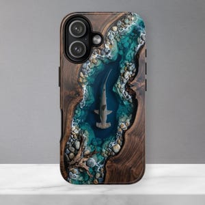 May include: A rectangular phone case featuring a wood grain border surrounding a central resin inlay. The inlay depicts a hammerhead shark swimming in deep blue water, bordered by light blue shallow water, white foam, and grey river stones.