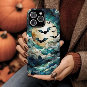 May include: A smartphone case featuring a Halloween design. The case displays a full moon with bats flying in front of it, set against a backdrop of blue and teal stained glass-style waves. The case has a black border and a camera cutout.