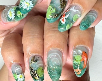 May include: Close-up of a hand with several oval-shaped nails painted with a green and white water-like design. Each nail features a different design, including a white flower, a gold fish, and a red and white fish.