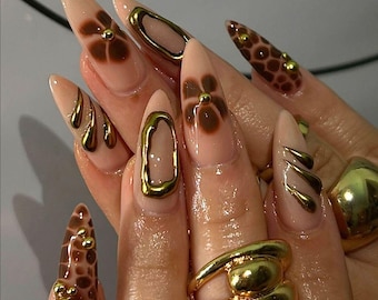 Puede incluir: Primer plano de una mano con uñas largas y almendradas pintadas de un color nude claro. Las uñas están decoradas con diseños geométricos dorados y marrones, incluyendo flores, remolinos y formas abstractas.